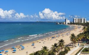 beautiful beach view with puffy clouds, clear ocean, and beach with palm trees and swimmers and city skyline in the background